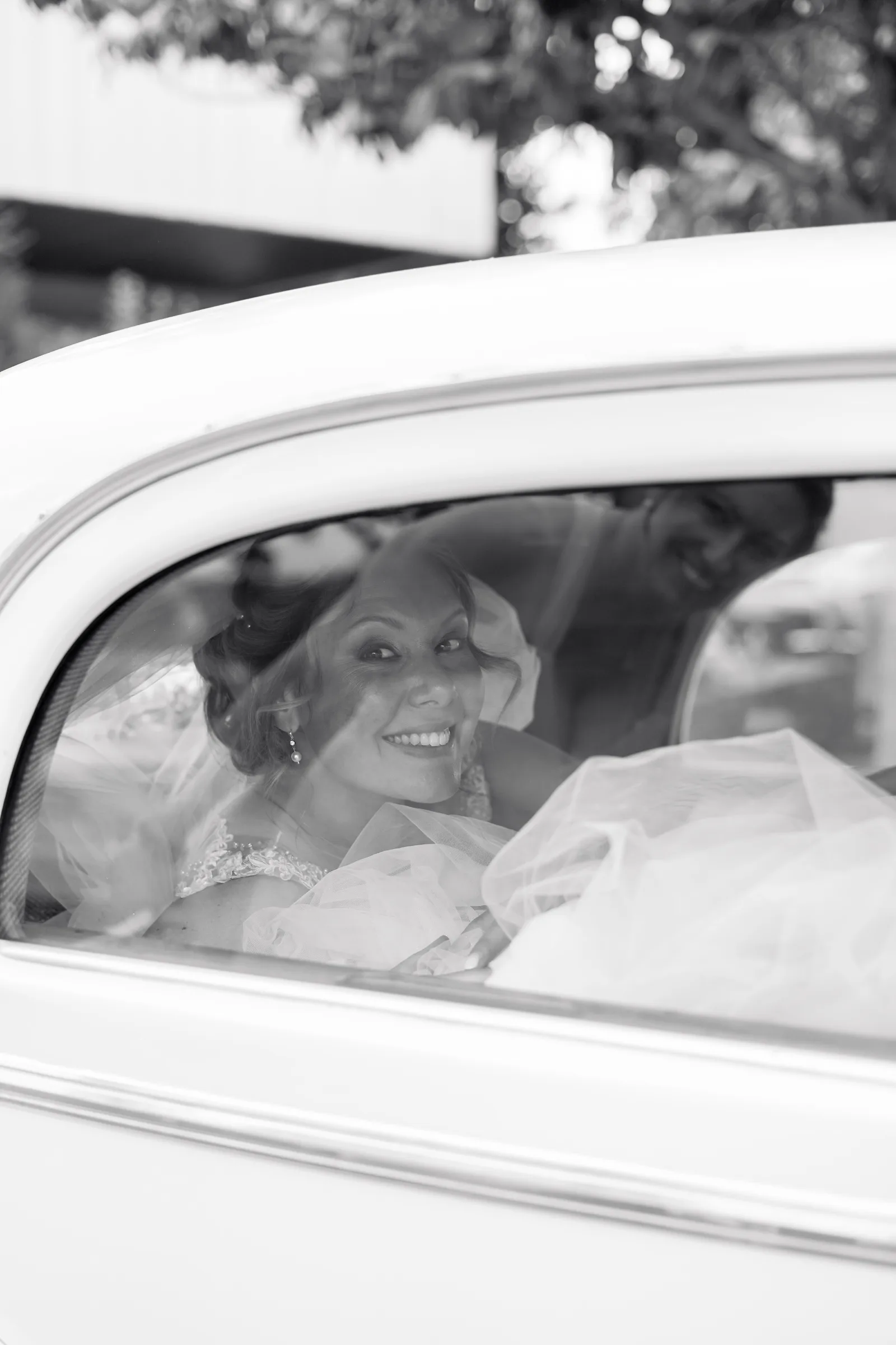 La mariée sourit derrière la vitre d’une voiture ancienne, en noir et blanc