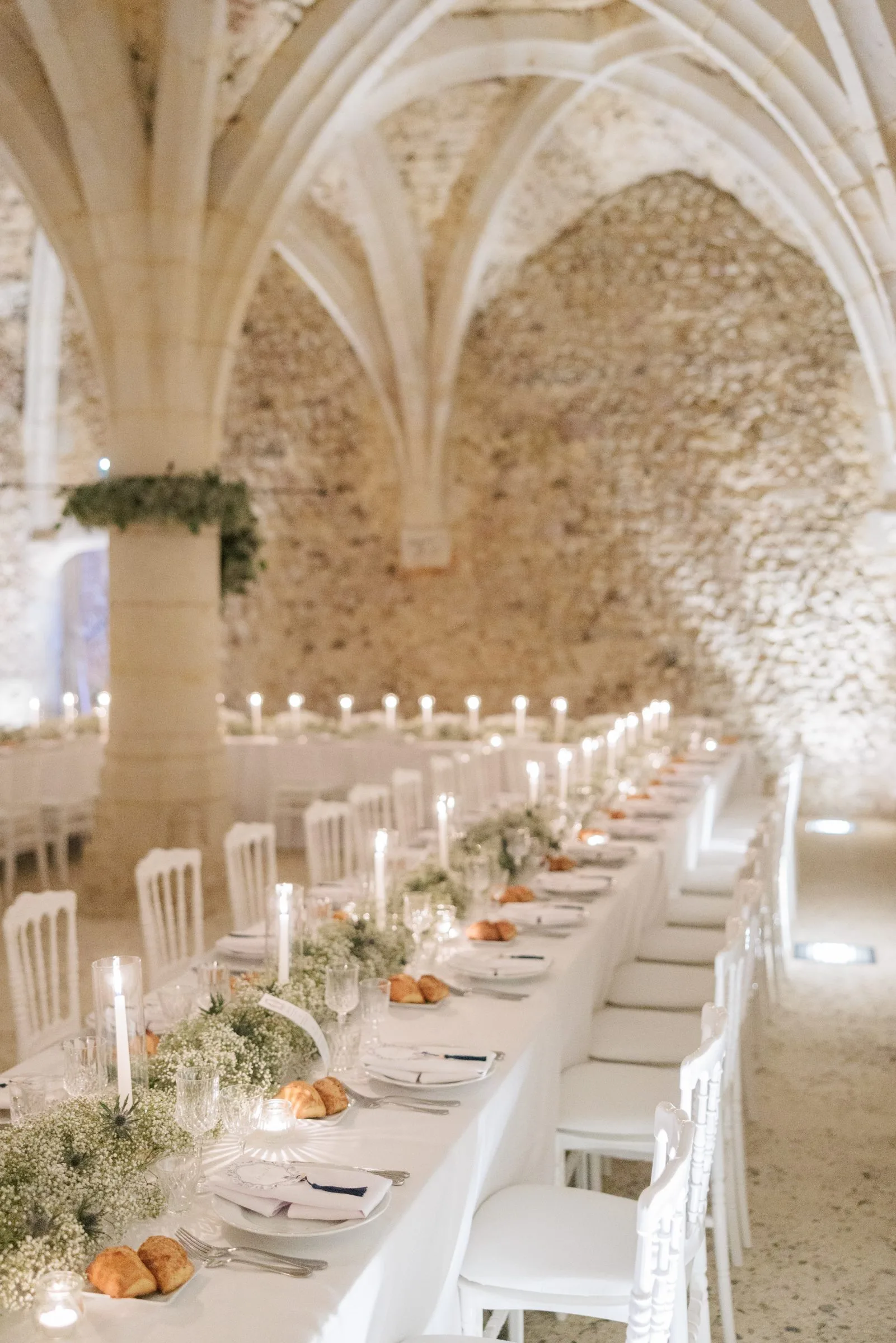 Longue table nappée de blanc sous les voûtes de pierre, bougies et gypsophile