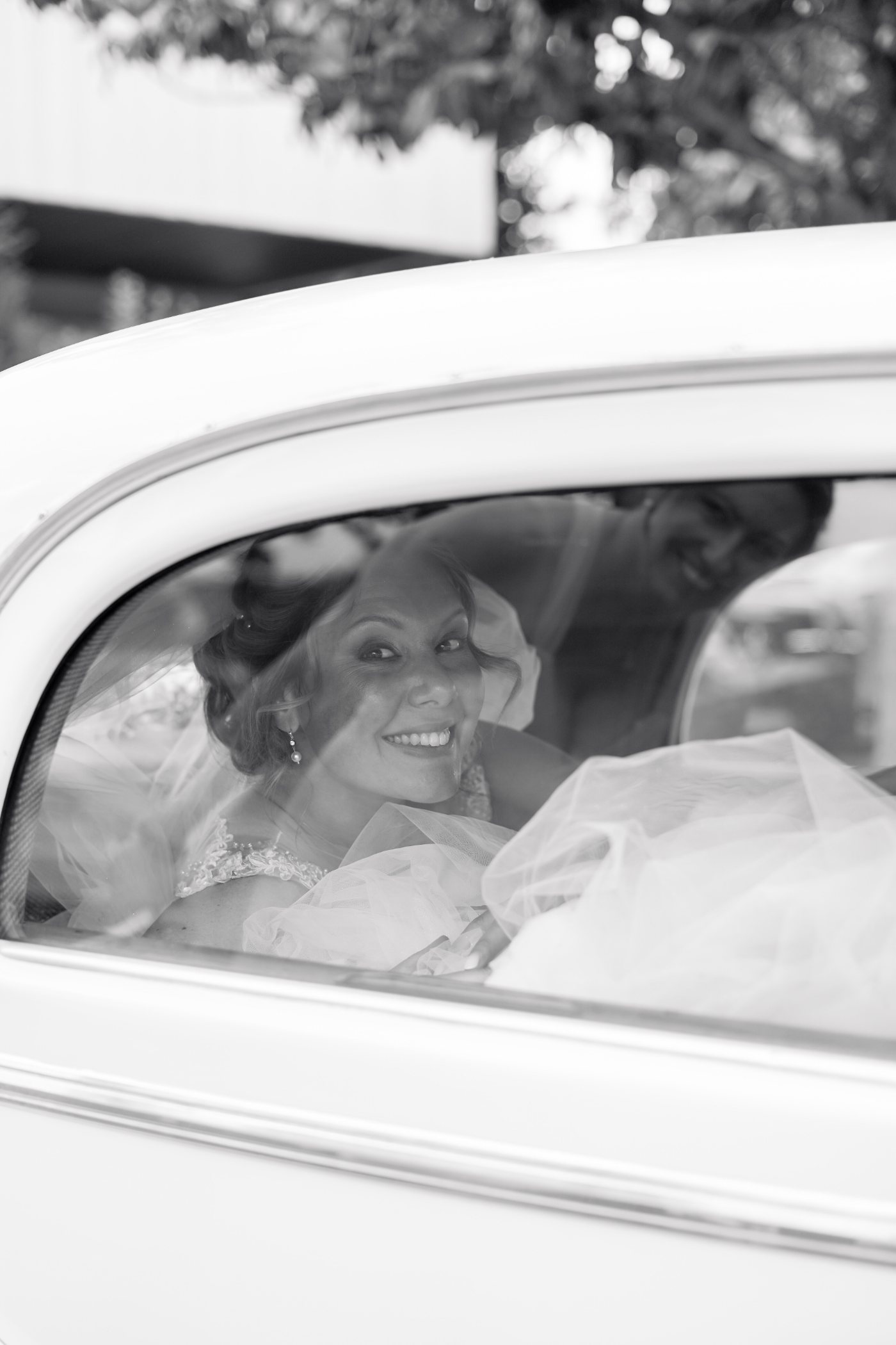 La mariée sourit derrière la vitre d’une voiture ancienne, en noir et blanc
