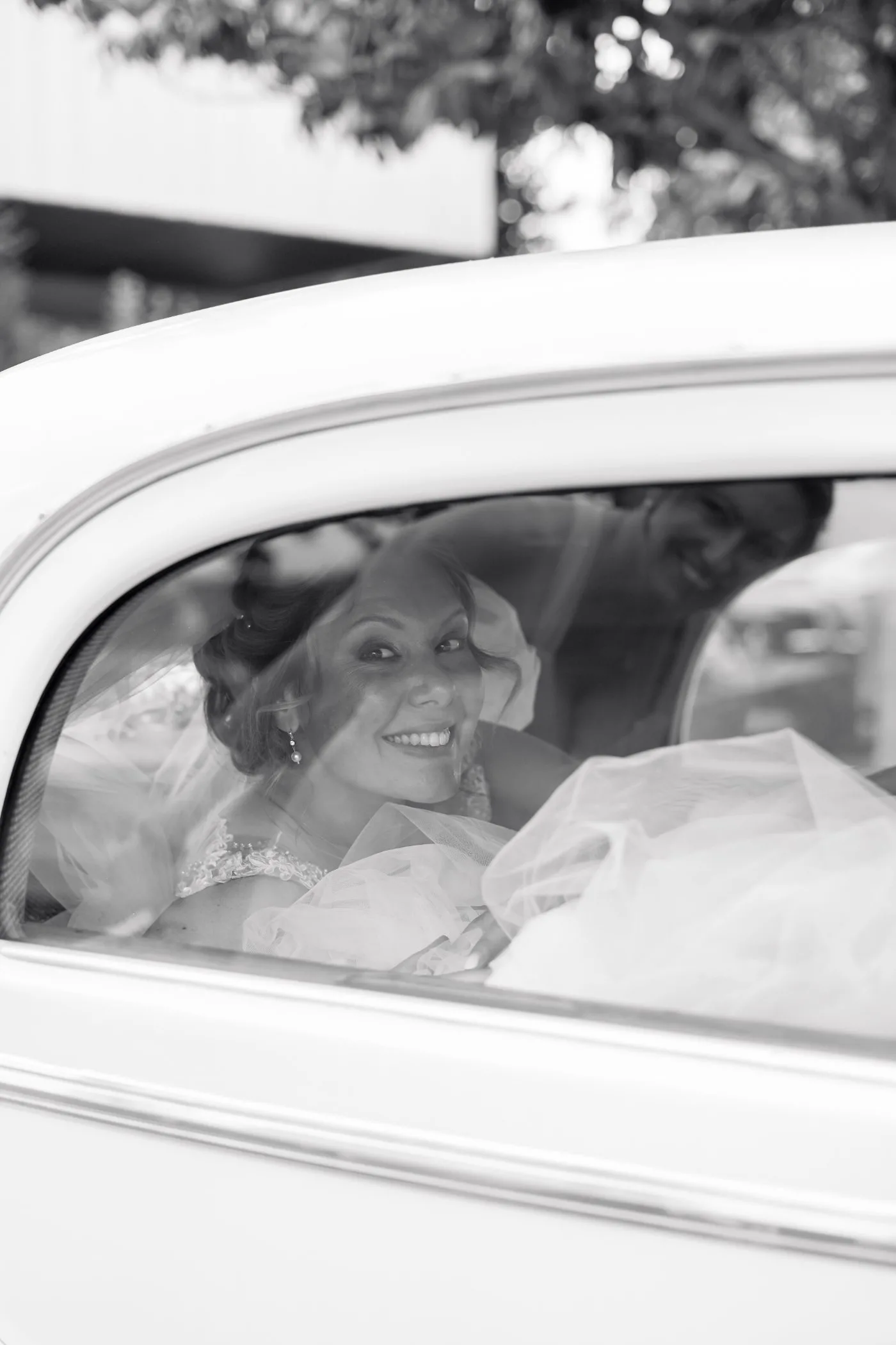 La mariée sourit derrière la vitre d’une voiture ancienne, en noir et blanc
